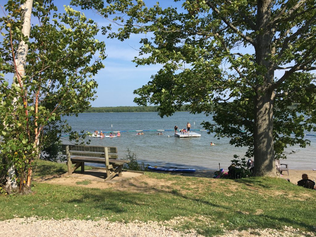 view from a bench looking out on to Miller Lake with people using swimming area at Miller's Family Camp